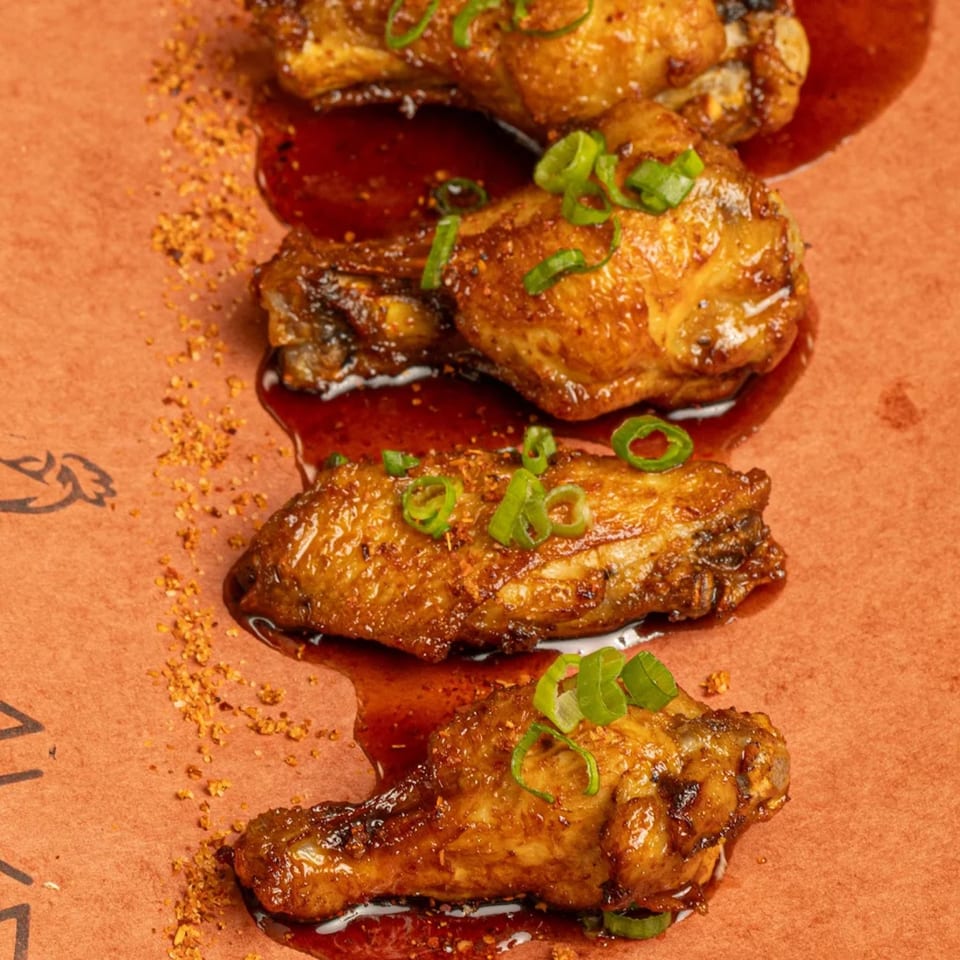 Four Peruvian chicken wings, glazed with sauce and garnished with green onions, on a brown surface.