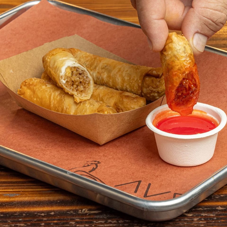 A hand dips a crispy Peruvian egg roll into a small cup of red sauce on a metal tray.